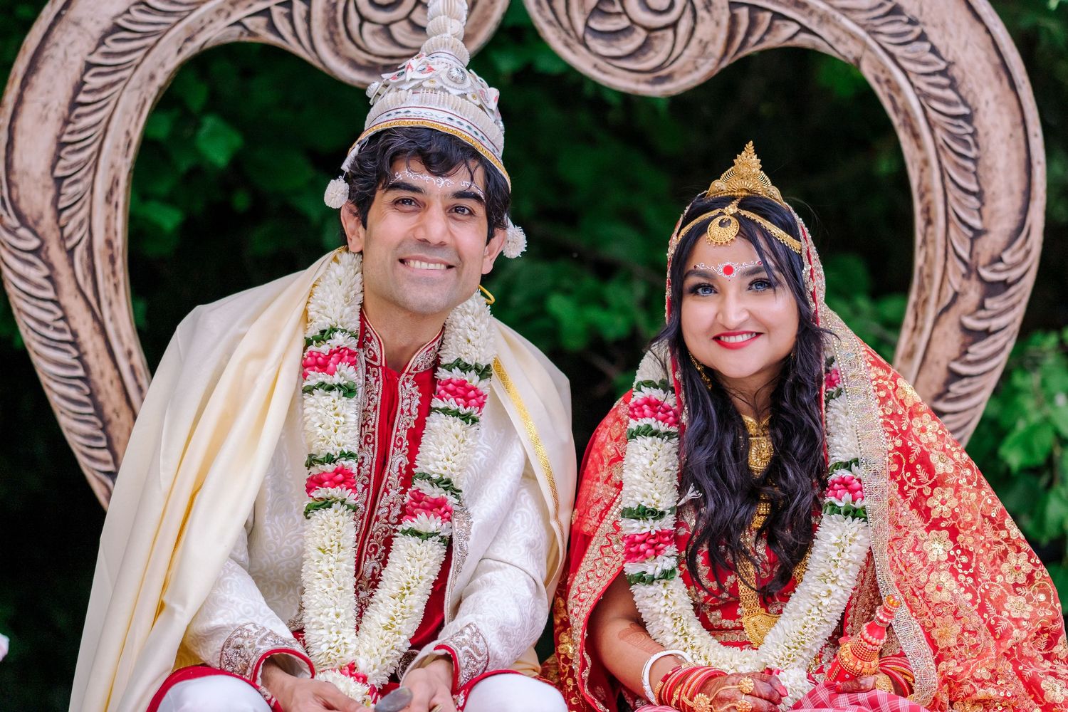 Traditional Indian wedding couple seated in ornate frame arch wearing ceremonial attire and garlands.