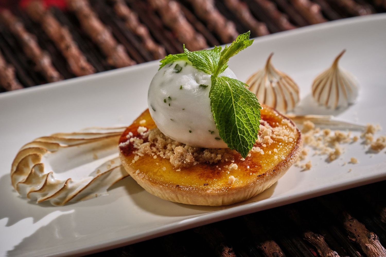 Elegant dessert tart topped with crème brûlée, whipped cream, fresh mint, and meringue kisses on a white plate.