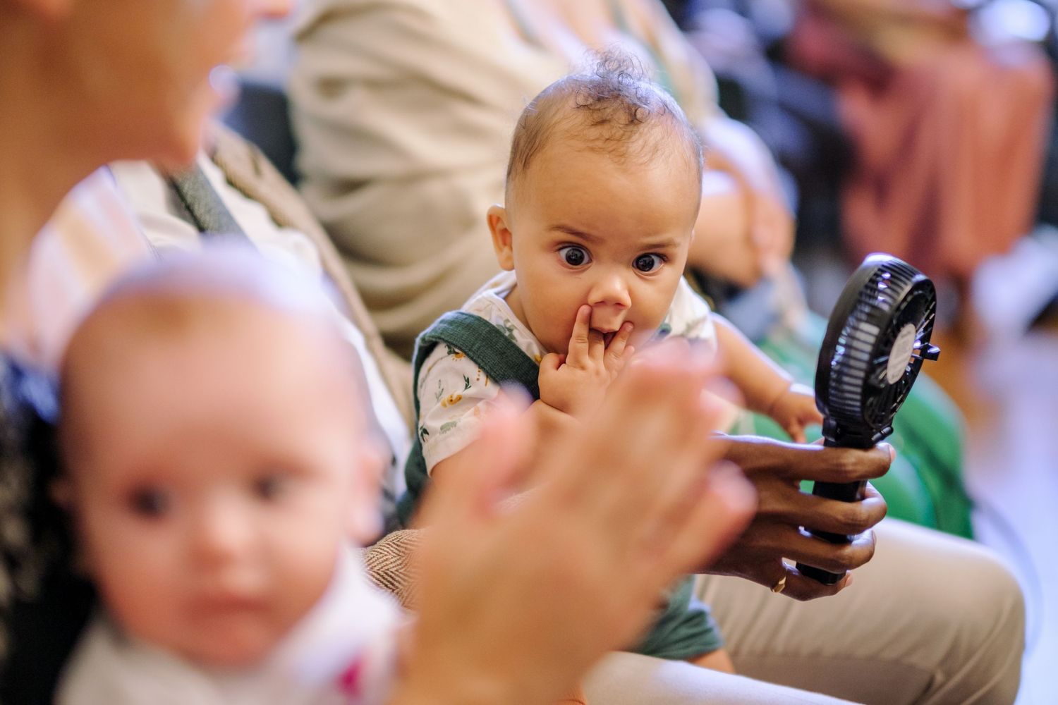 A small child sits among a crowd while someone holds a handheld fan nearby.