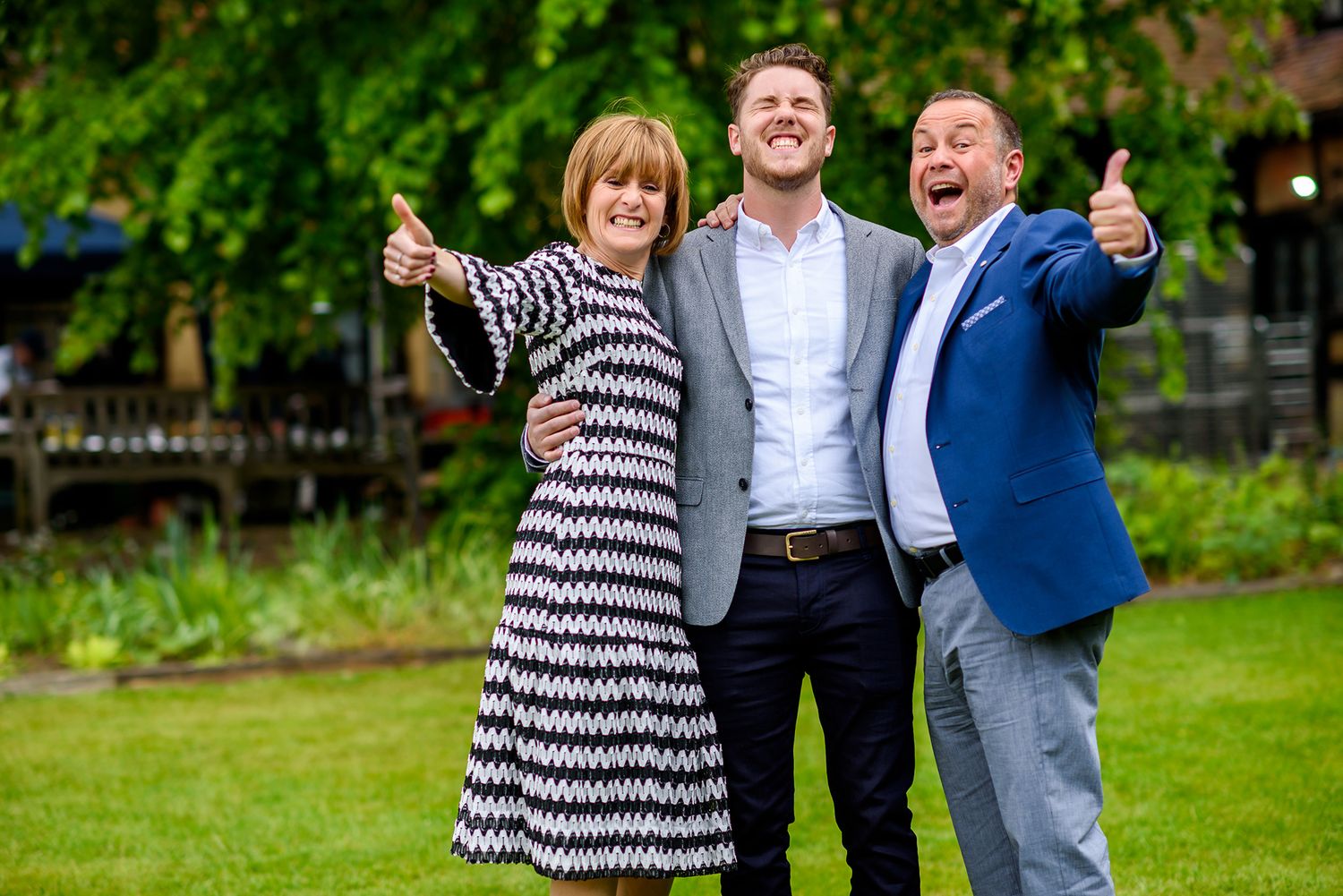 Three people pose cheerfully with thumbs up in a garden setting.