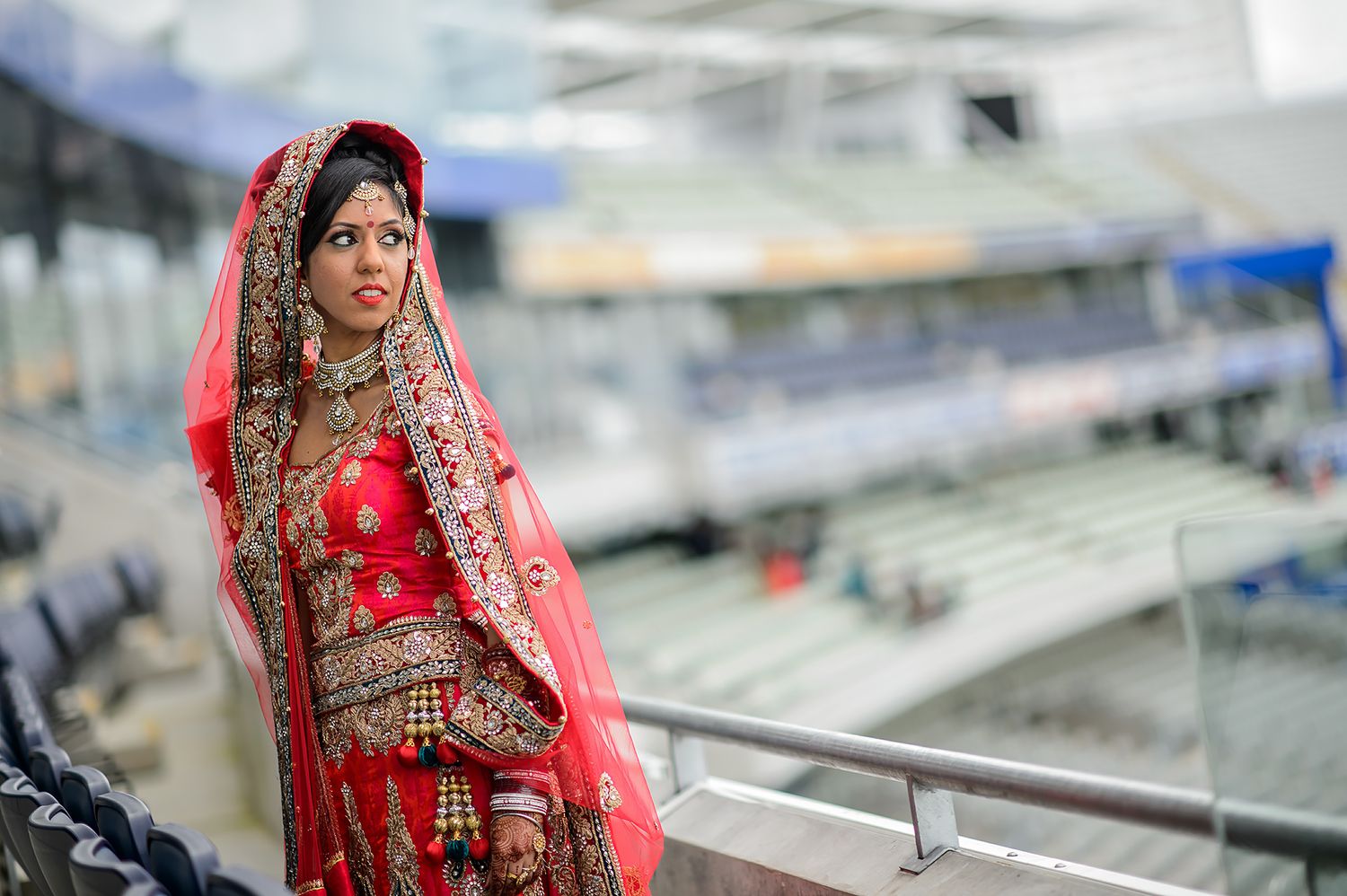 Couple in traditional Indian wedding attire posing against stadium backdrop.