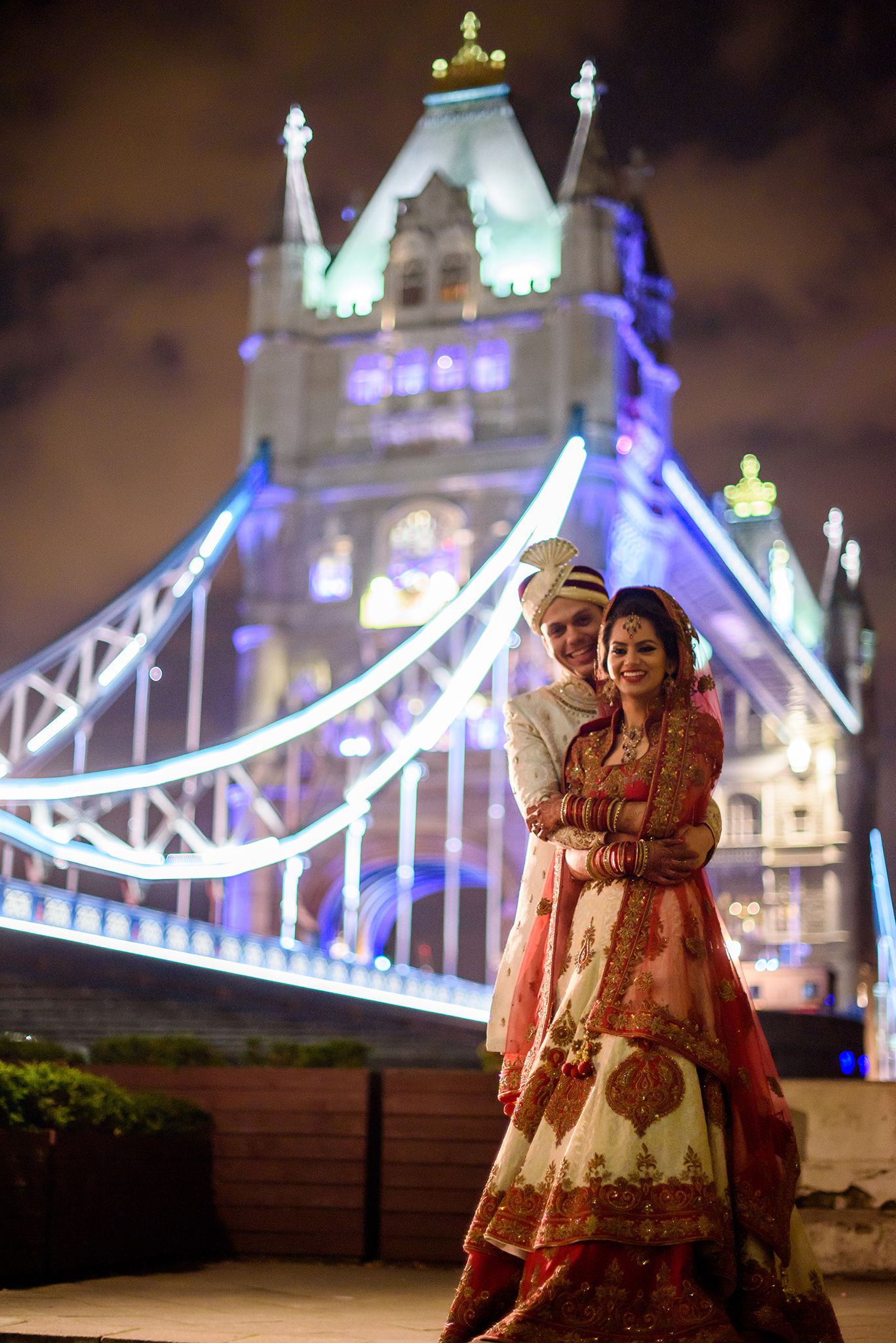 A couple in traditional Indian wedding attire pose before the illuminated Tower Bridge at night.