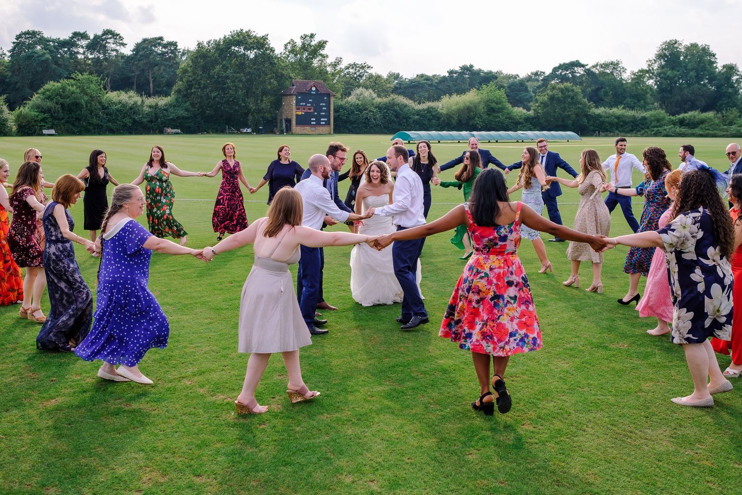 Wedding guests dancing in a line on grassy field during outdoor celebration.