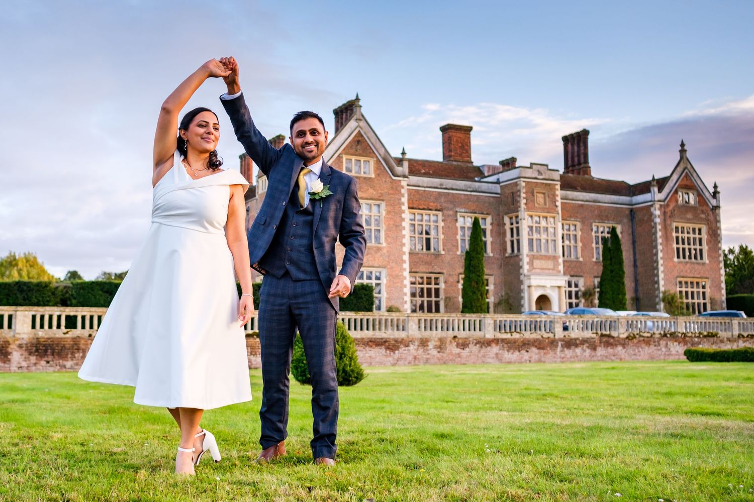 A couple in wedding attire walks on grass with a historic brick building in the background.