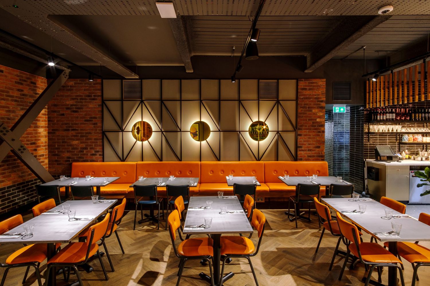 Modern restaurant interior with orange chairs and wooden tables illuminated by warm lighting and industrial ceiling.