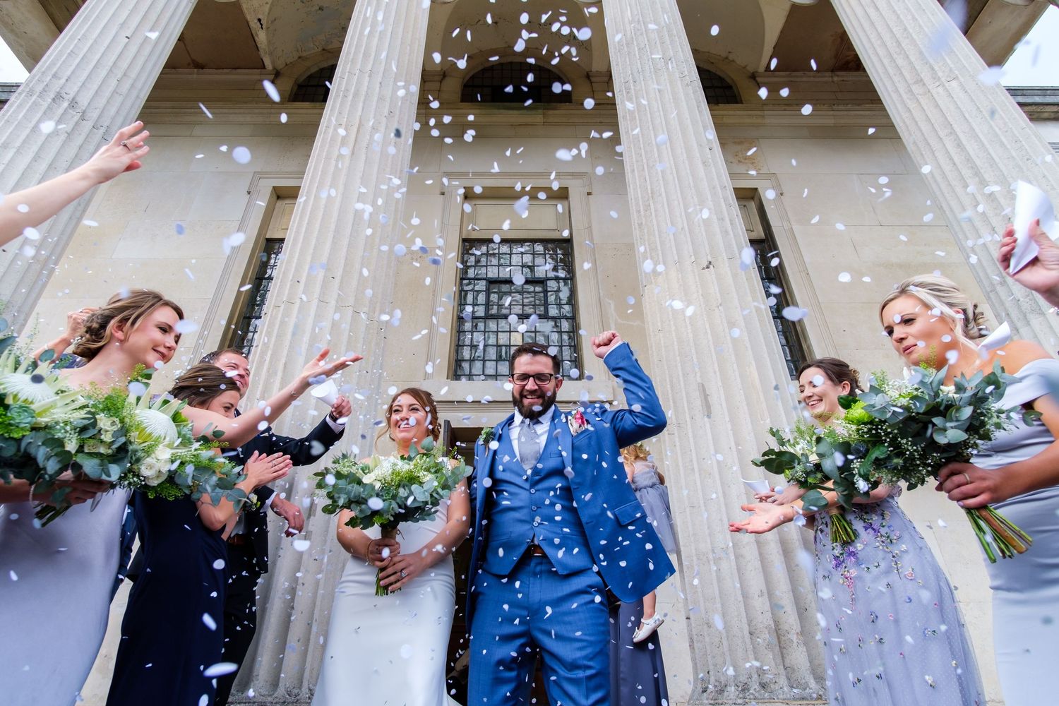 Wedding party celebrates with confetti outside a columned building entrance.