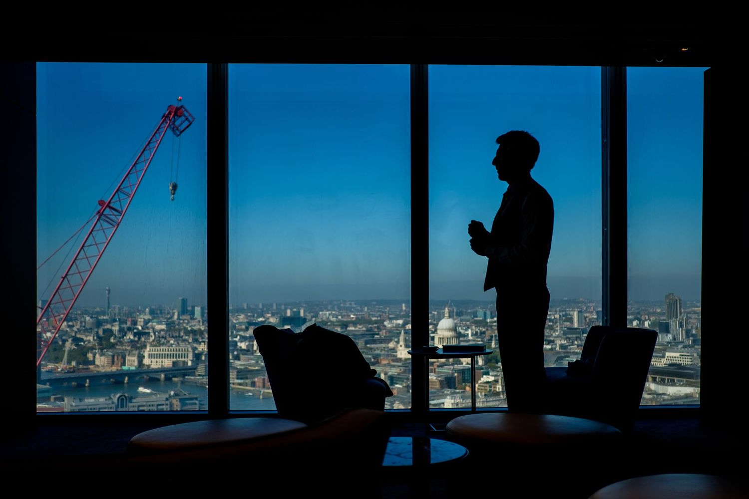 Silhouetted figures gaze at a cityscape through windows while construction cranes dot the blue sky outside.