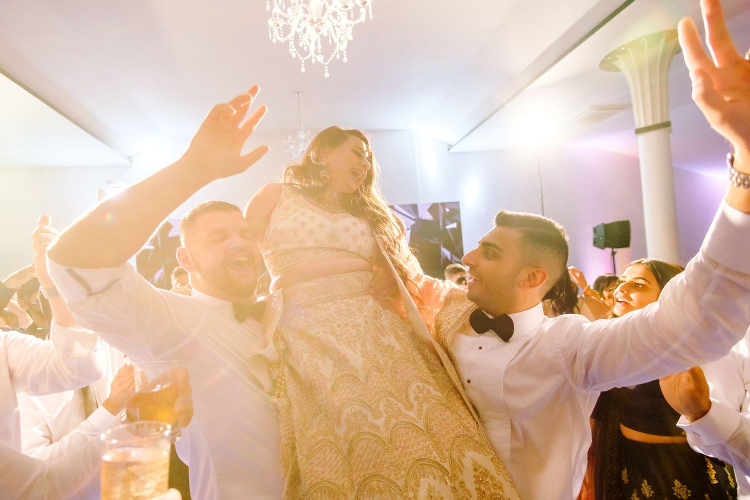 Joyful celebration with dancing and raised hands under crystal chandelier at wedding reception.
