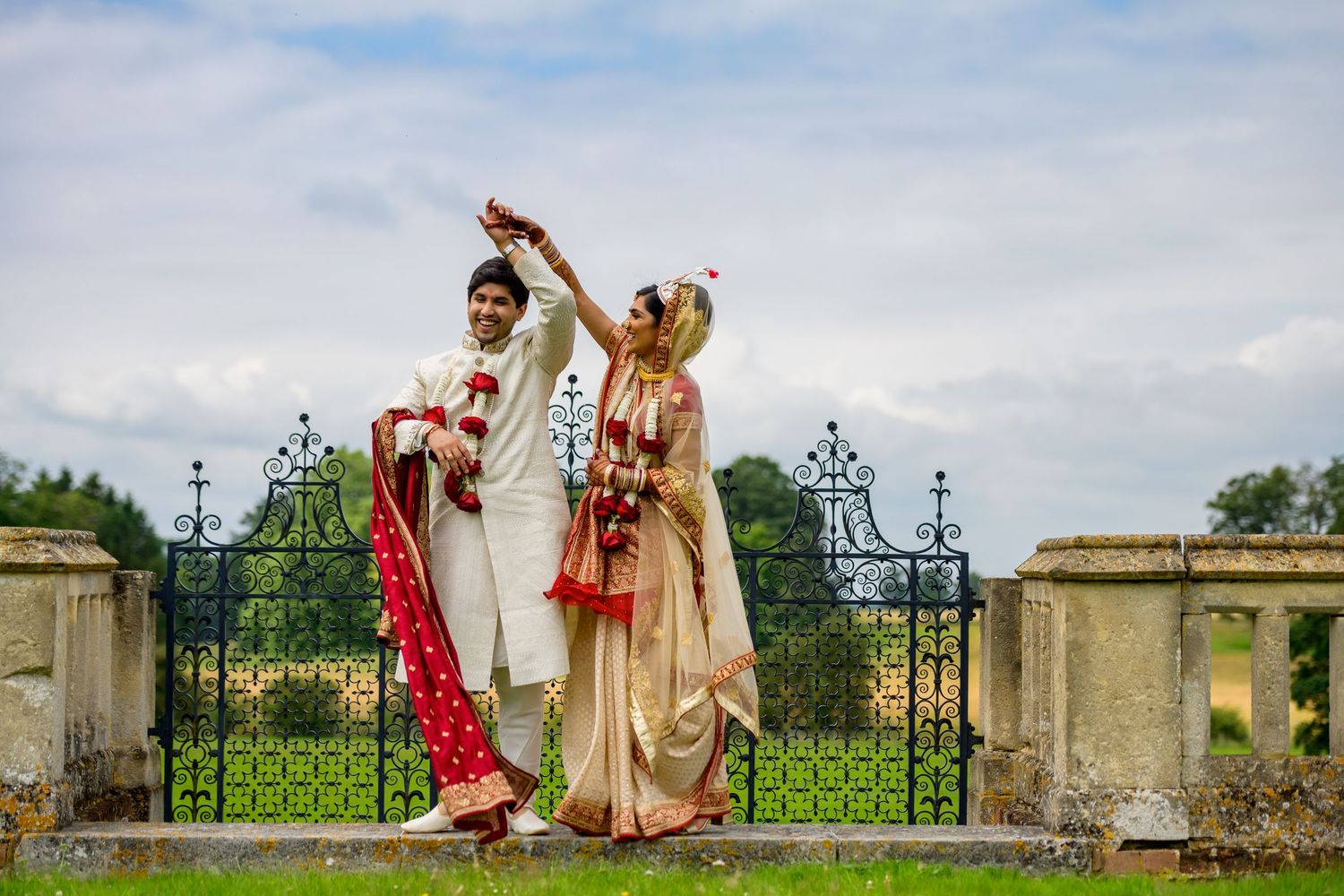 Wedding party members dance together outdoors at an elegant garden venue with ornate gates and greenery.