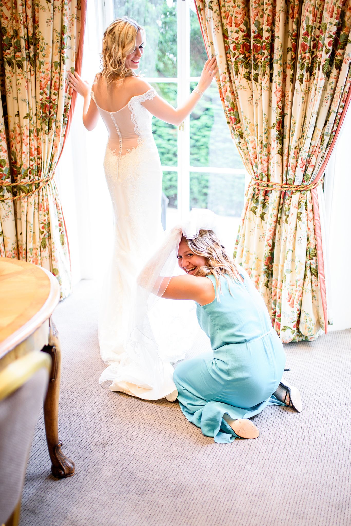 Bridesmaids in turquoise dresses help with the wedding gown against floral curtains.