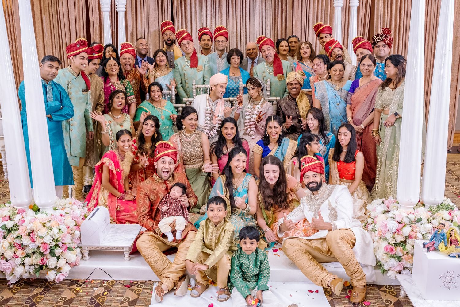 A large group of wedding guests in colorful traditional Indian attire pose together for a celebratory photo.