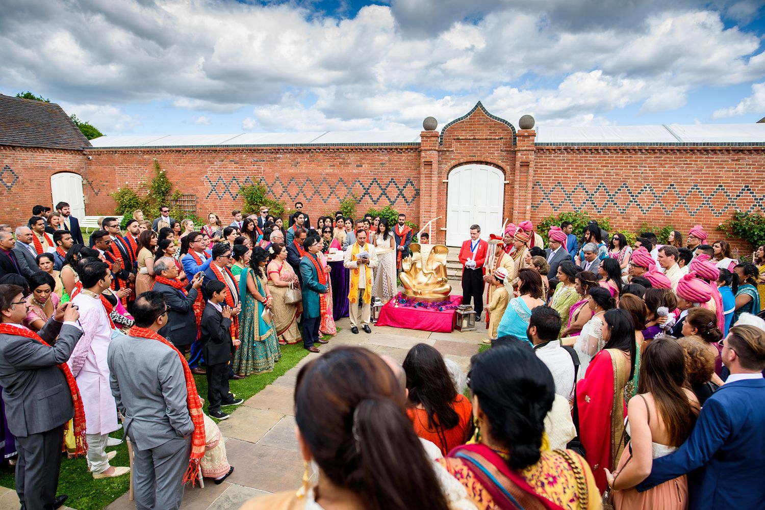 Large wedding gathering in front of ornate brick wall with archway during outdoor ceremony.