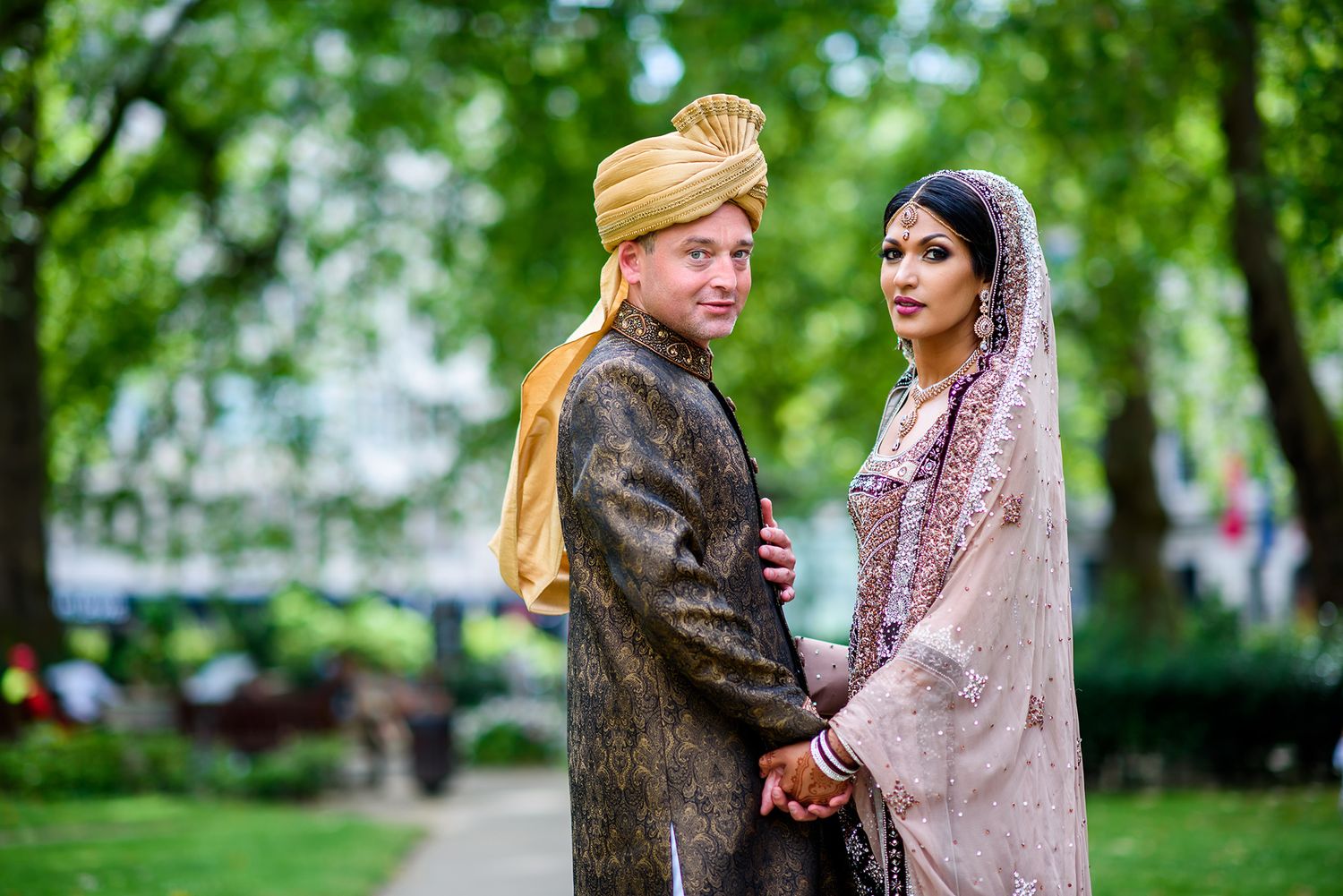 Couple in traditional South Asian wedding attire embracing in a lush green park.