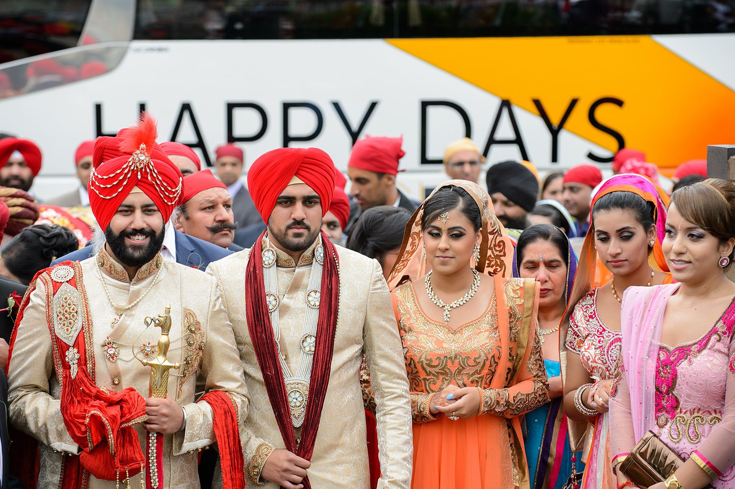 Traditional Indian wedding party poses in front of a 'Happy Days' sign in ornate ceremonial attire.