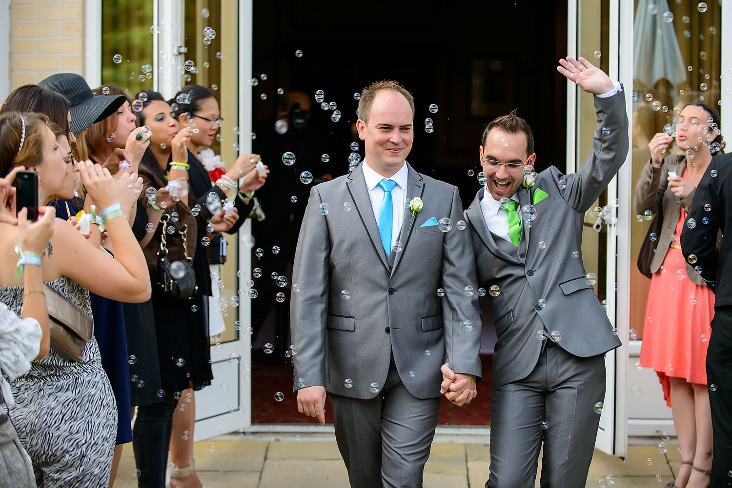 Celebration with bubbles as couple exits venue in matching gray suits with turquoise accents.