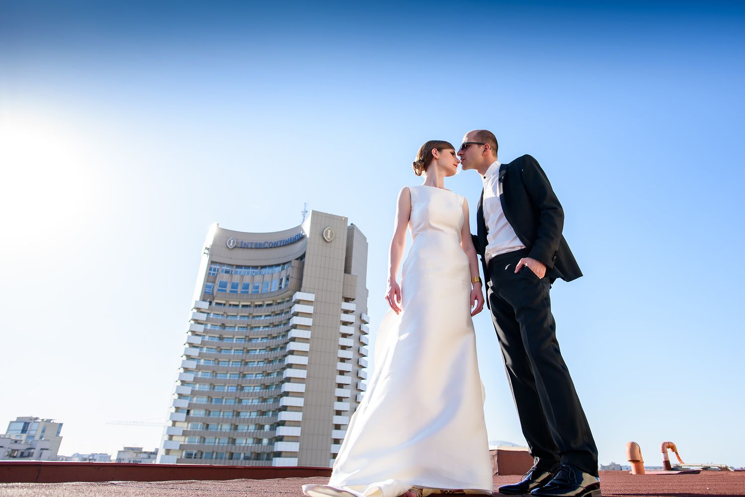 A formal couple embraces outdoors with a modern high-rise building visible in the background.
