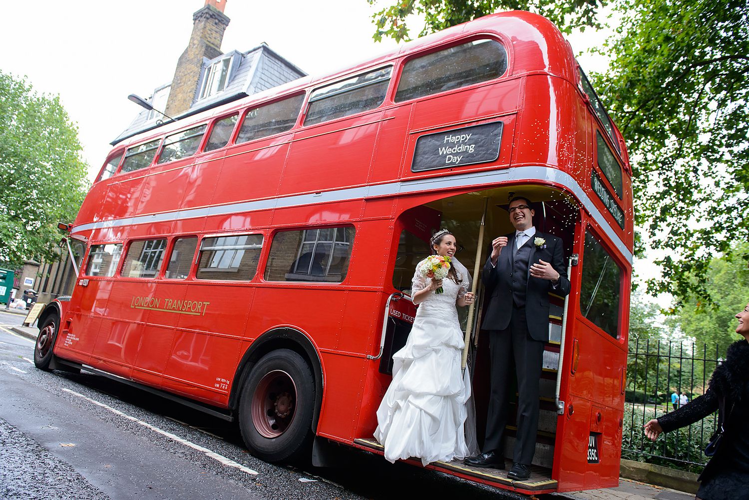 Classic red double decker bus serves as unique wedding transportation on tree-lined street.