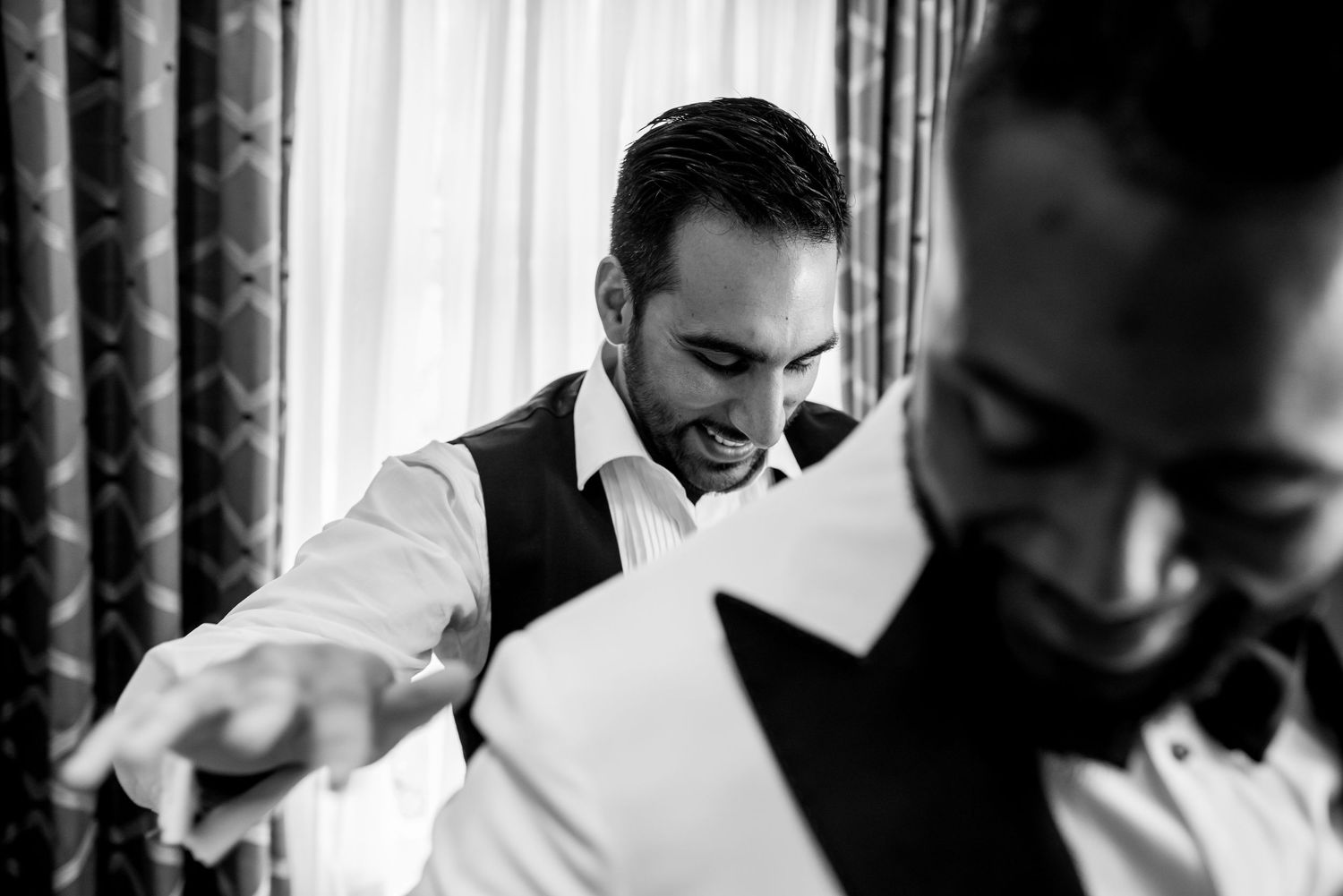 A candid black and white photo showing wedding preparations with the groom getting ready in formal attire.
