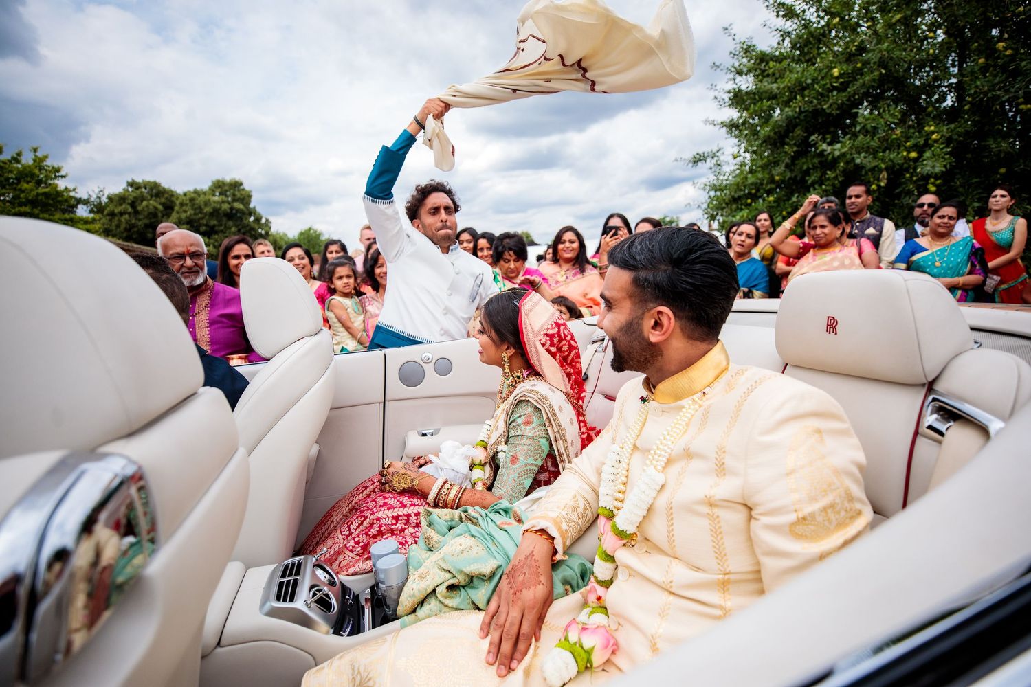 A wedding celebration shows people enjoying festivities from a vintage white convertible car outdoors on a sunny day.