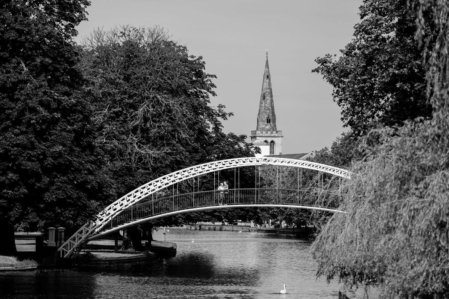 Black and white photograph of an arched bridge over water with a church spire in the background.