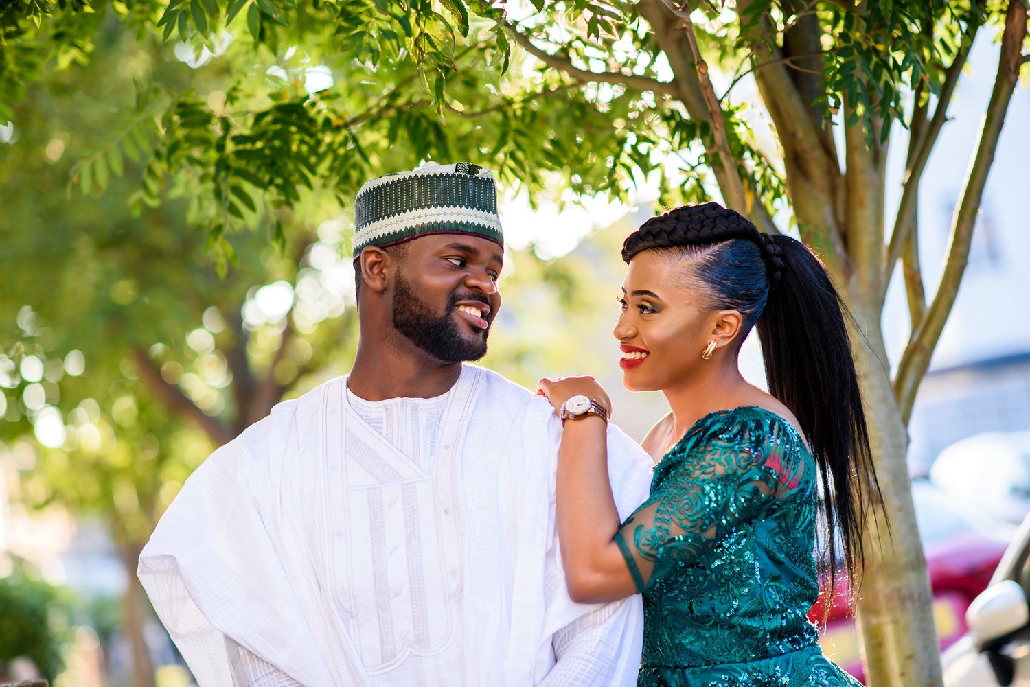 A romantic outdoor photoshoot features a couple in white and teal outfits under leafy trees.