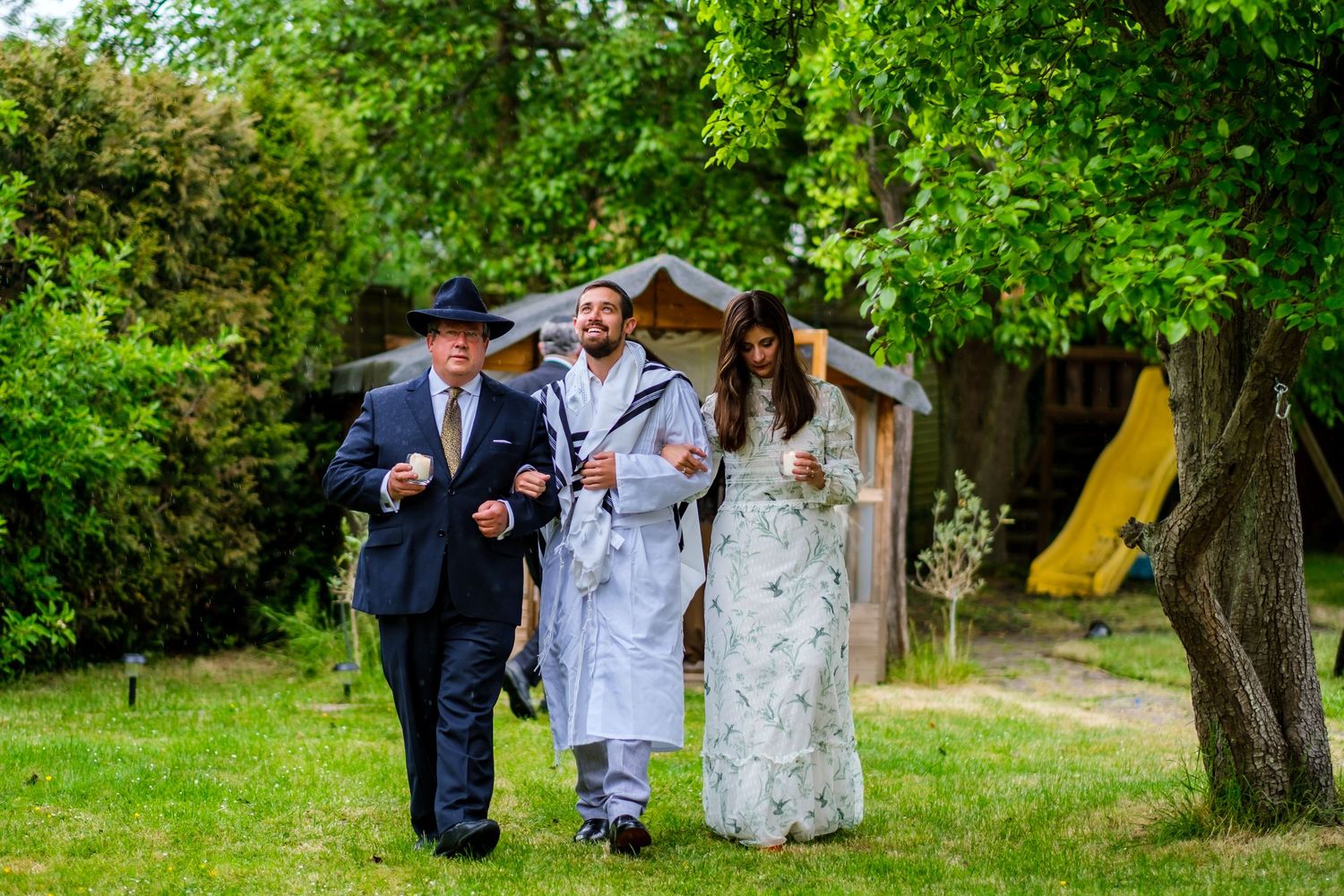 Wedding party walks together through a lush green garden setting during an outdoor celebration.