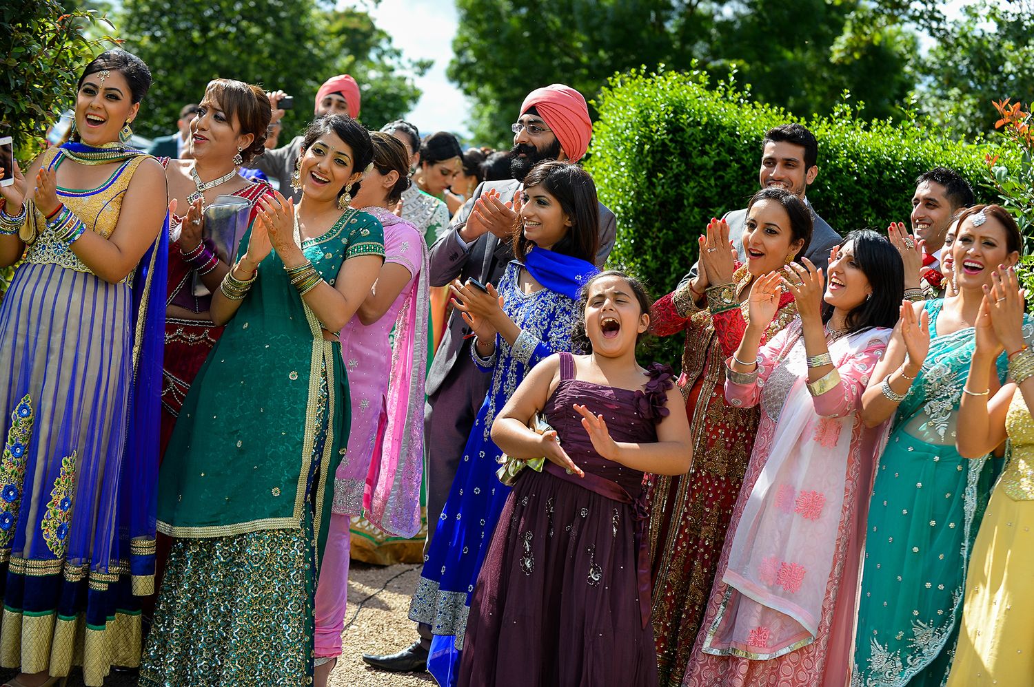 Group of wedding guests in colorful Indian attire cheering outdoors.