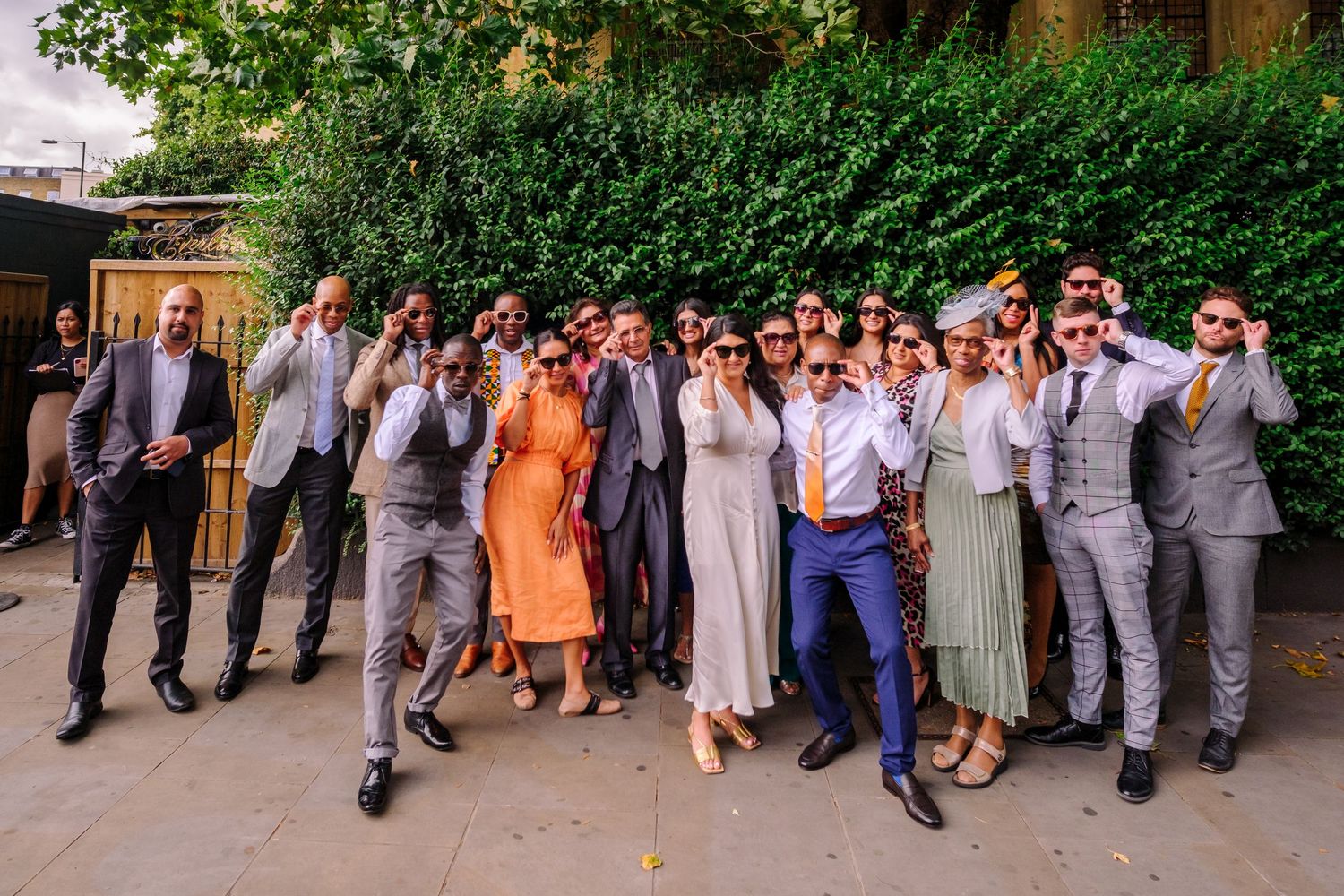 A large wedding party poses together making surprised expressions in a garden setting.