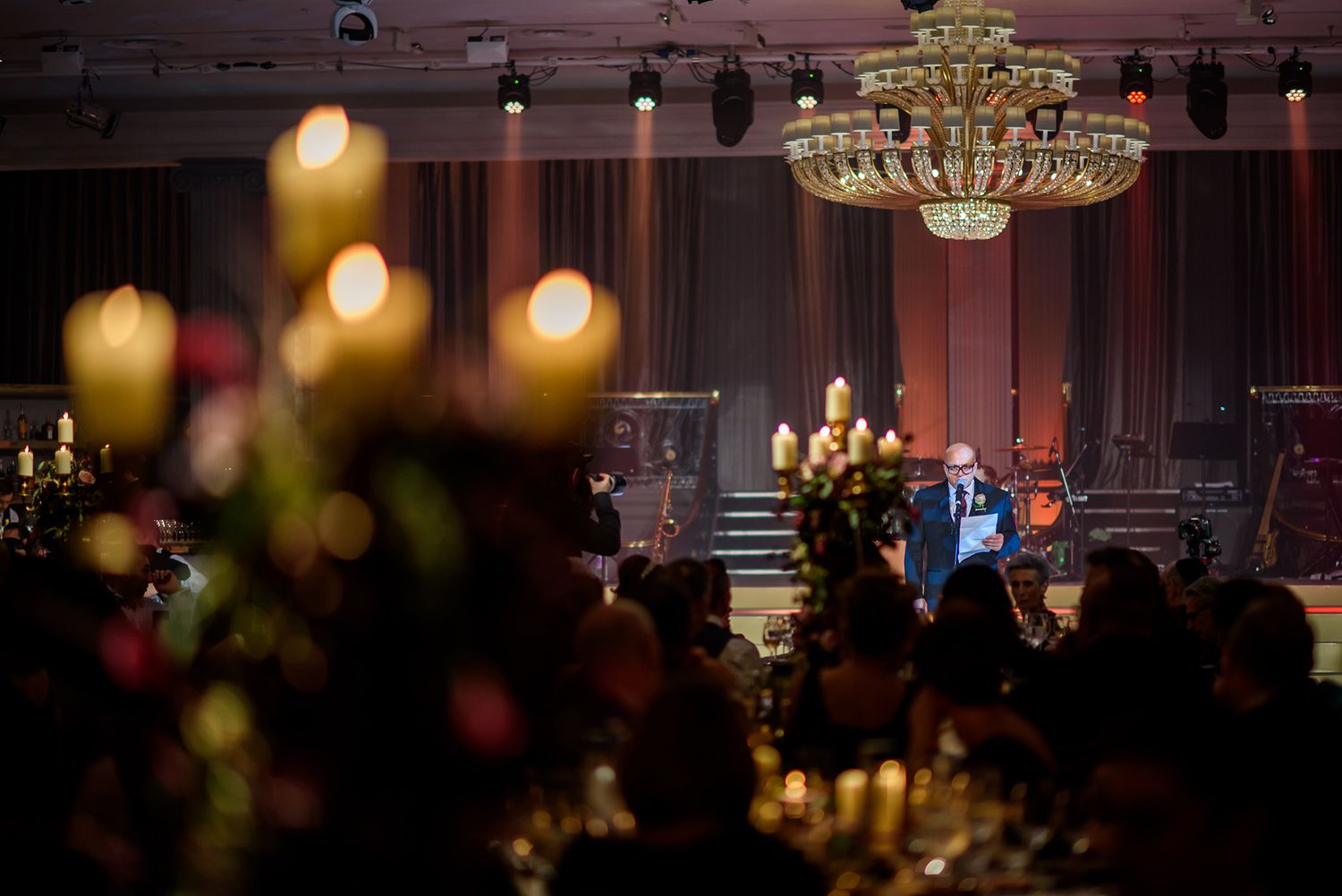 Candlelit wedding reception with crystal chandelier and stage in romantic dark ambiance.