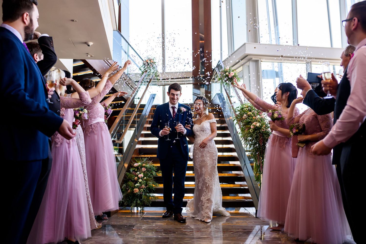 A wedding party stands on wooden stairs while exchanging bouquets and flowers during an indoor ceremony.