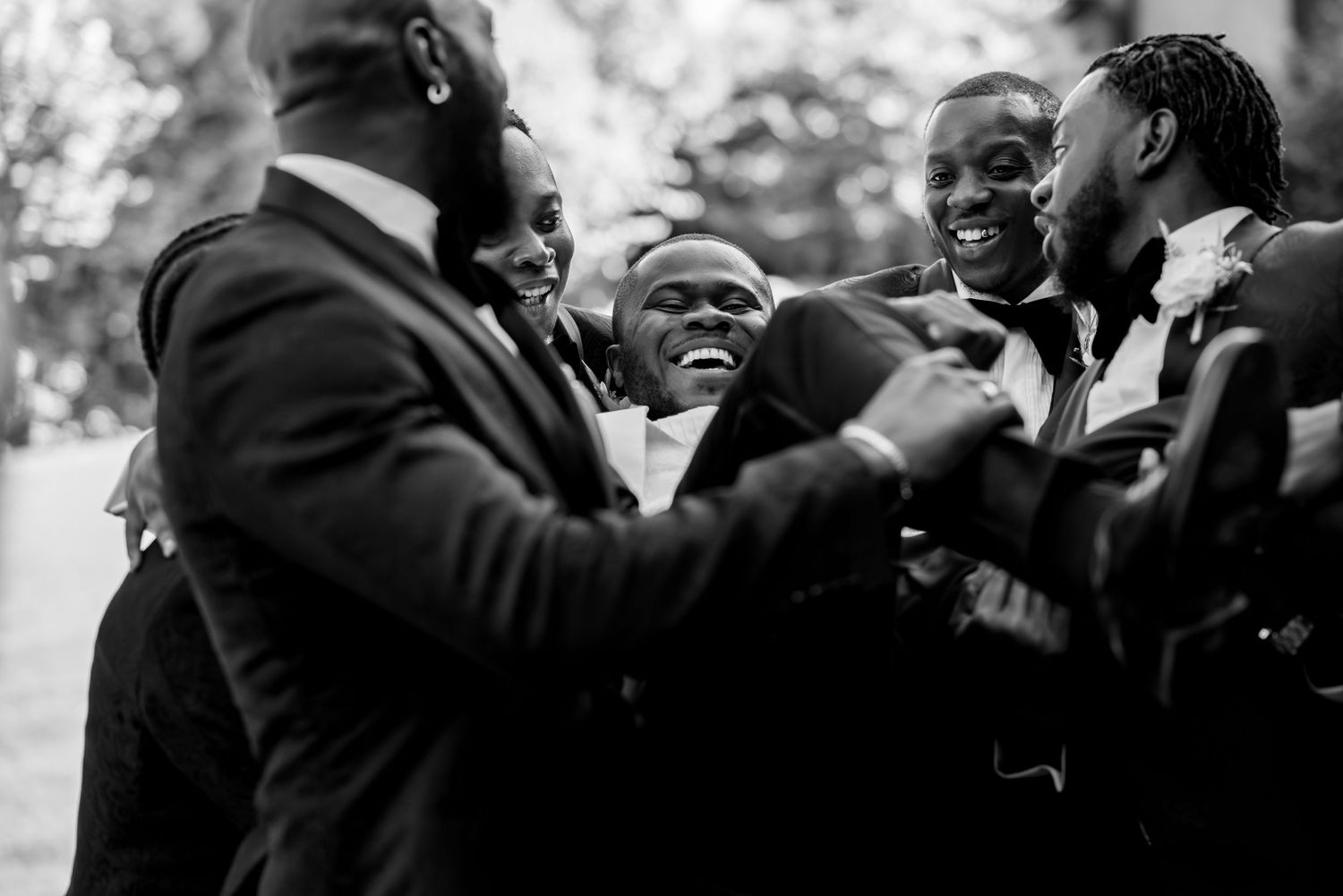 A candid black and white photo captures joyful laughter among groomsmen at an outdoor wedding.