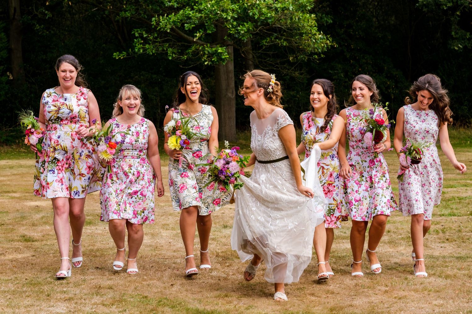 Bridesmaids in floral dresses running through a sunny field together.