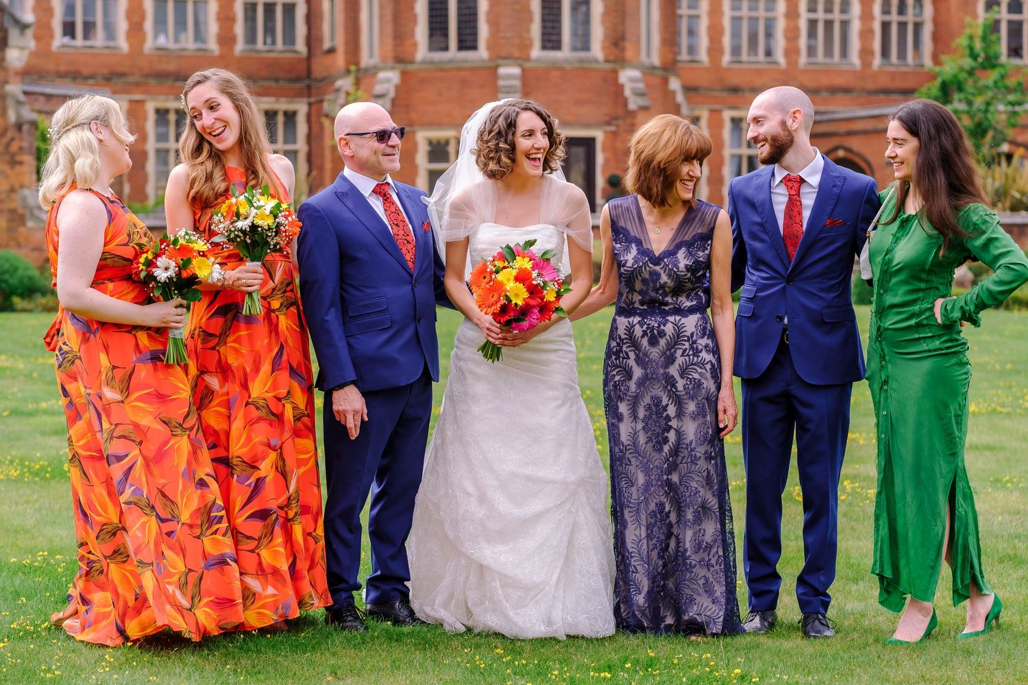 Wedding party poses on lawn wearing vibrant orange, navy blue and green attire with colorful fall bouquets.