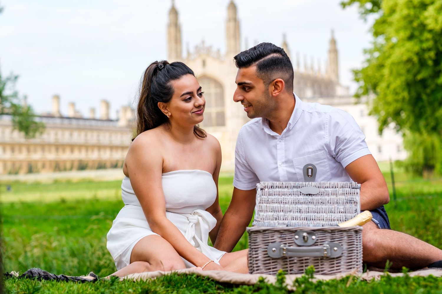 A couple relaxes on a picnic blanket in a lush green park with a historic building visible in the background.