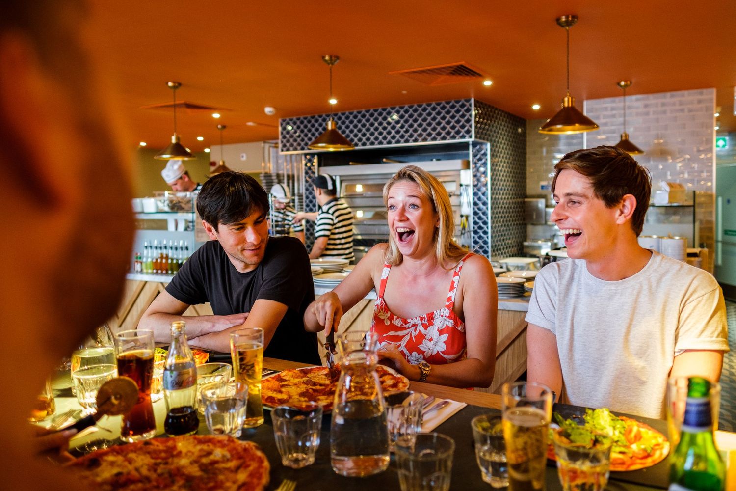 People gathered at a restaurant bar counter with pizzas and drinks in warm ambient lighting.