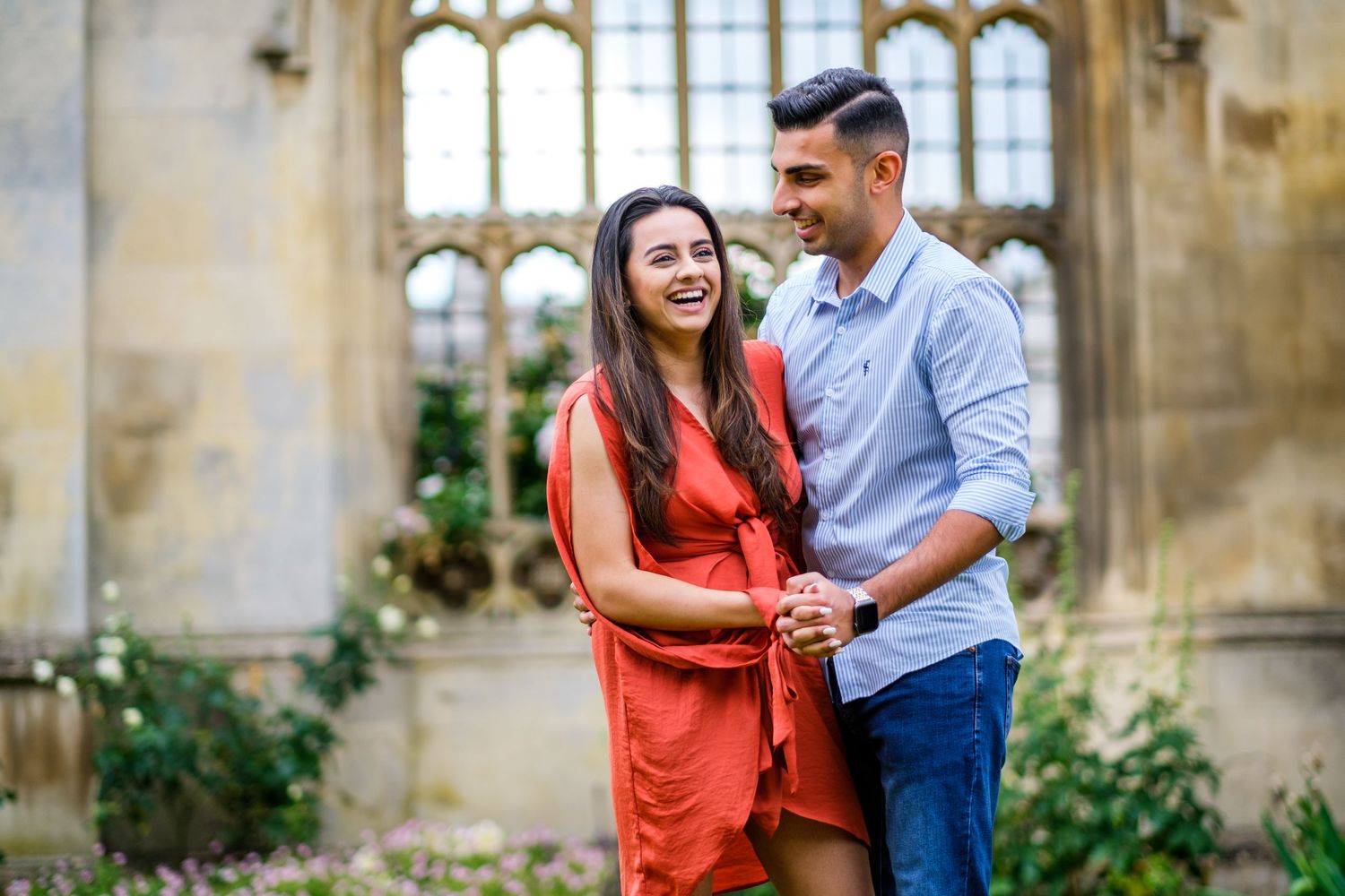 A couple shares a romantic moment in front of a historic stone building with Gothic windows and climbing vines.