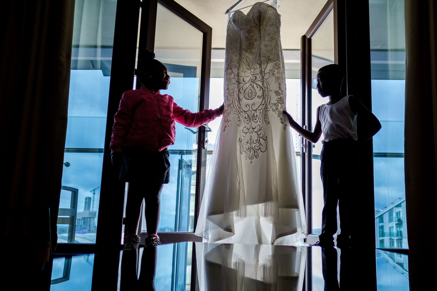 Silhouette of someone holding up a white wedding dress near a window with city views in the background.
