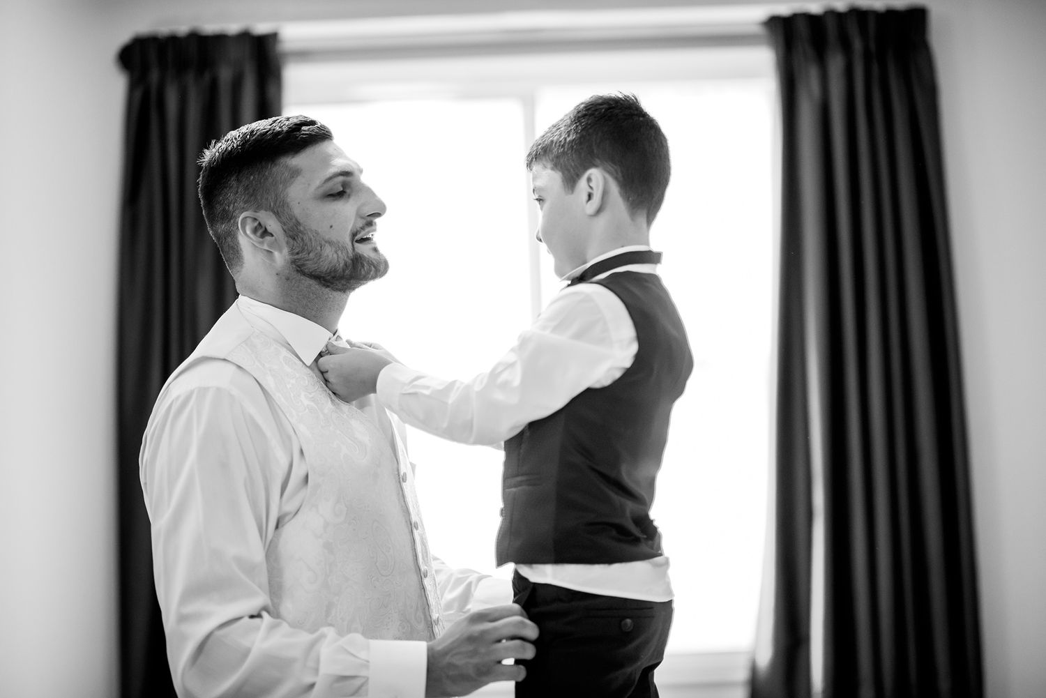 A tender black and white moment of formal clothing adjustments by a window.