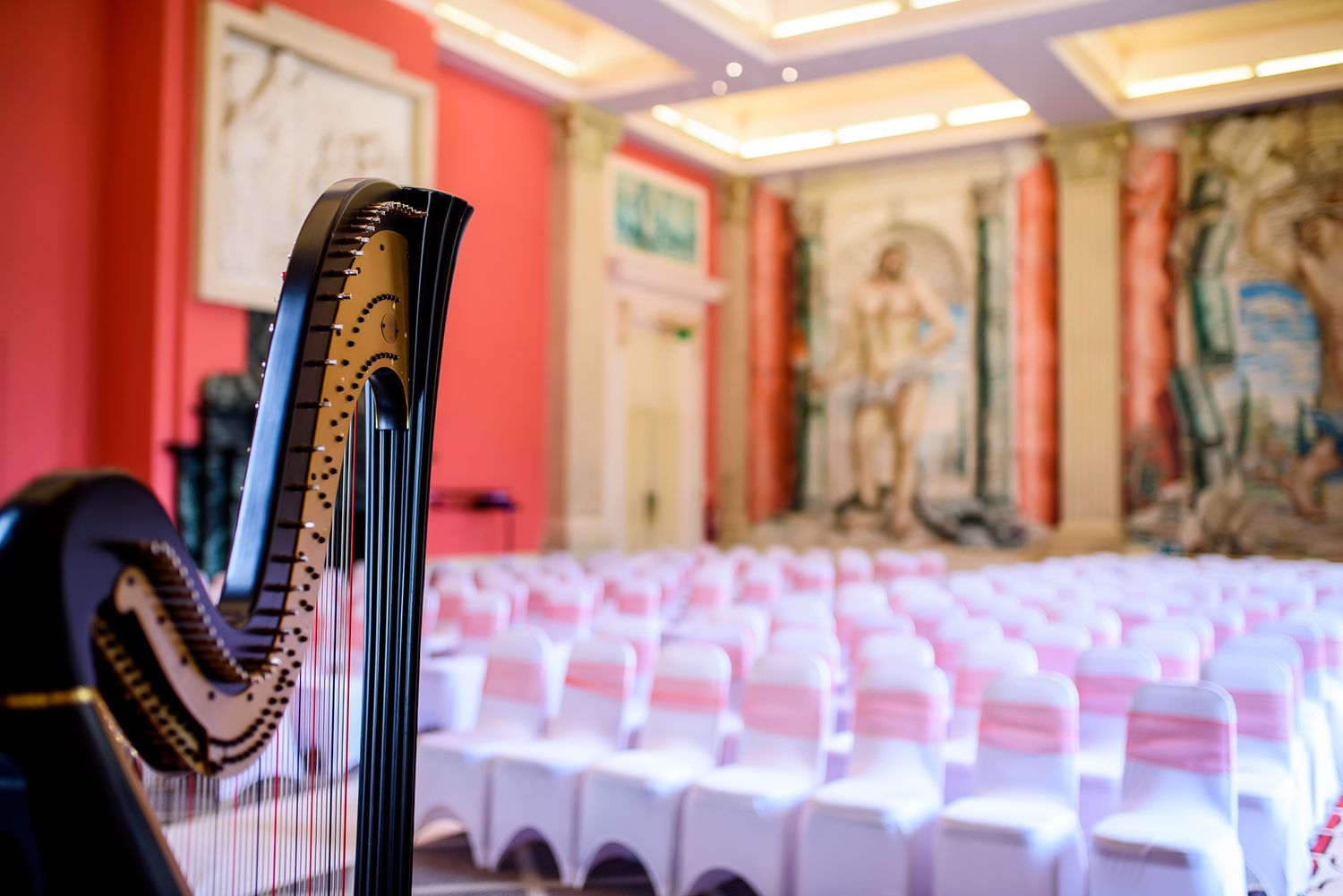 Elegant harp beside rows of white chairs in ornate concert hall with decorated walls.