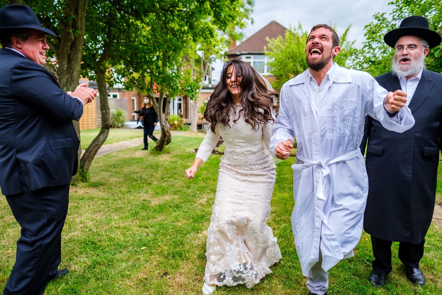 A joyful wedding celebration with attendees dancing together on a green lawn next to buildings.