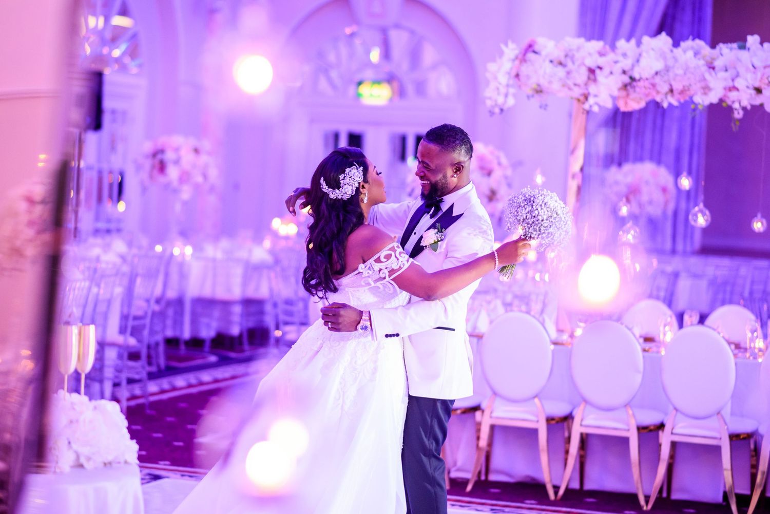 Elegant purple-lit wedding reception with white decor and chandeliers.