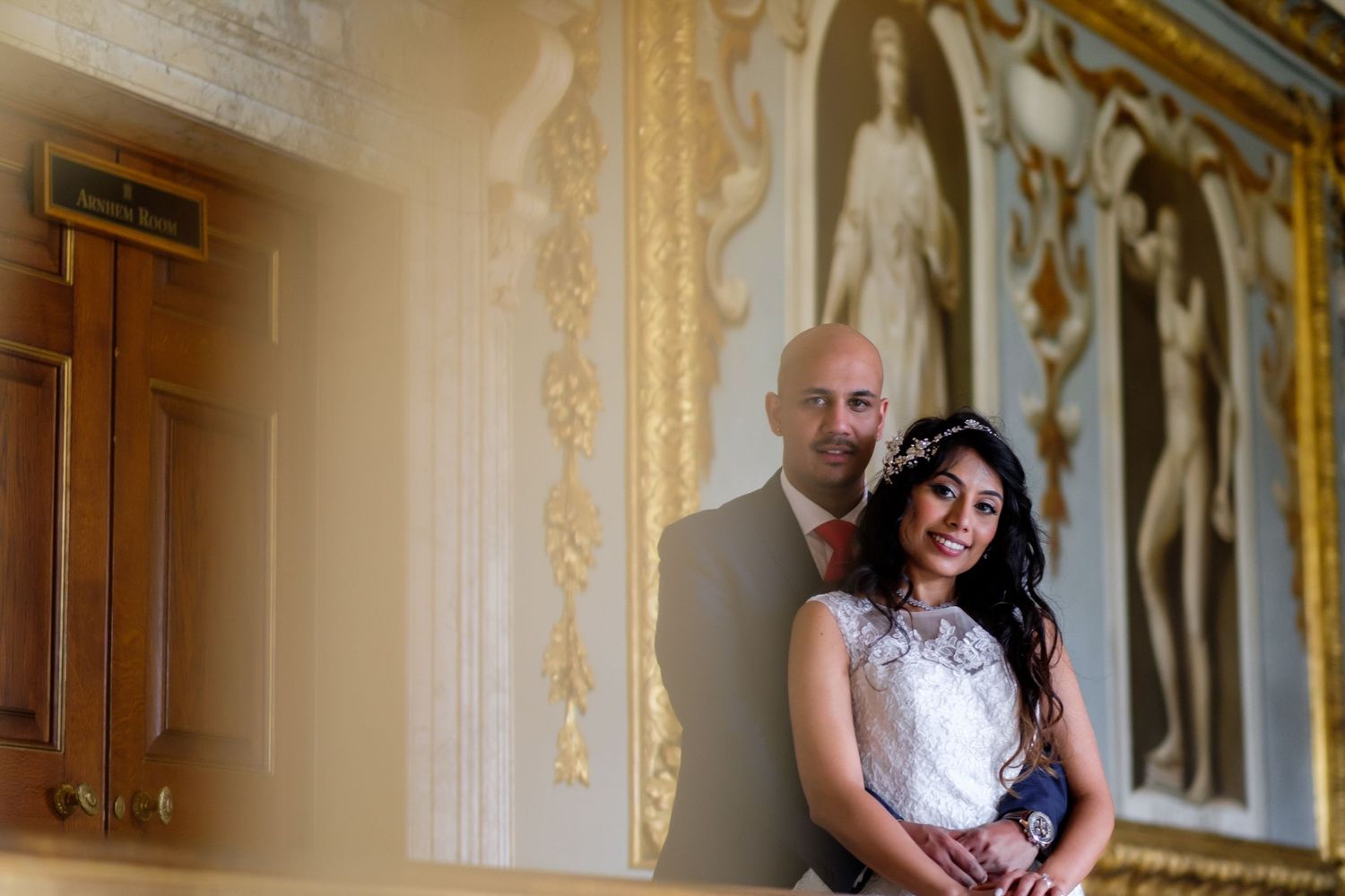Wedding portrait against an elegant cream-colored wall with ornate gold details.