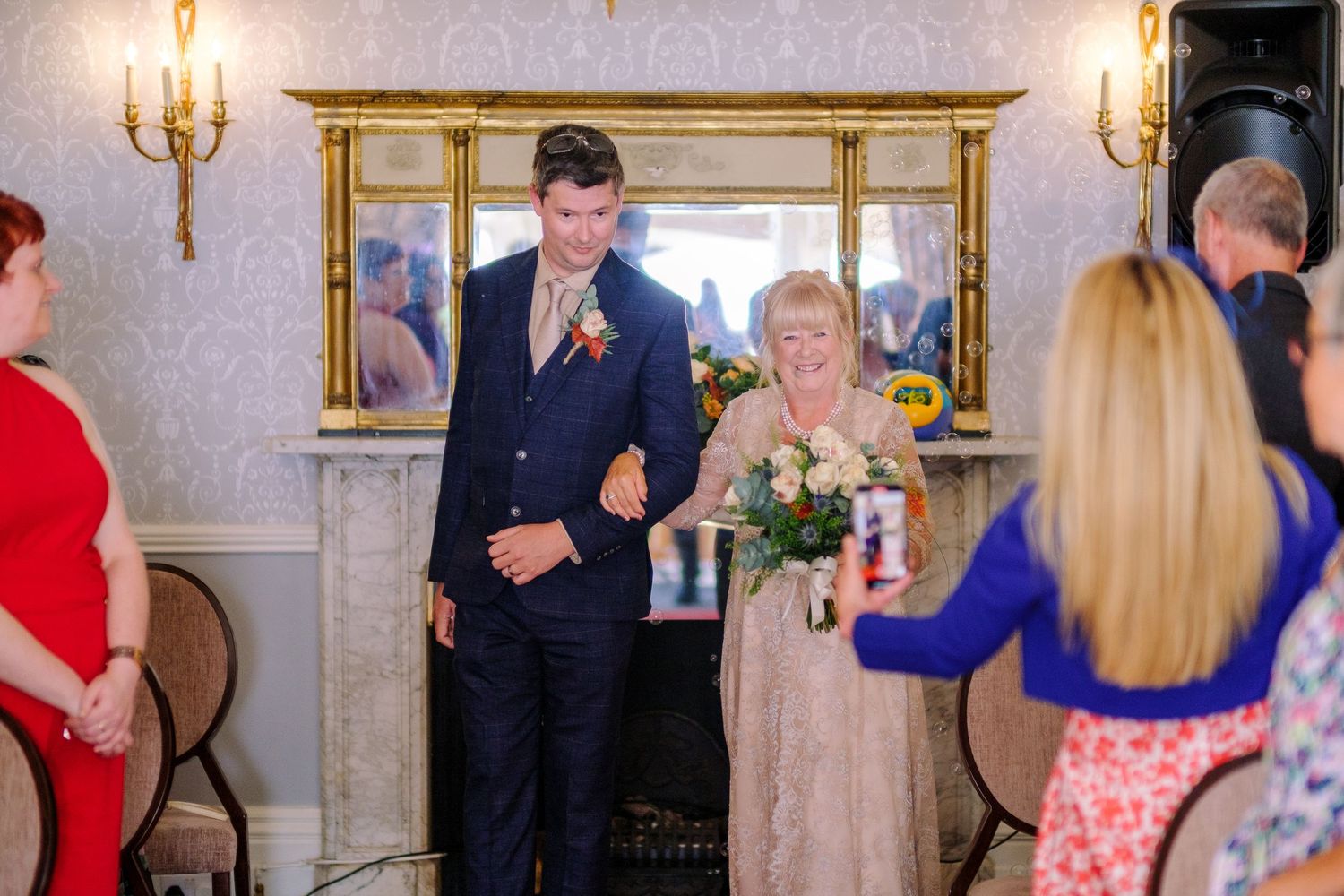 Wedding reception scene with guests mingling under elegant wall sconces and chandeliers.