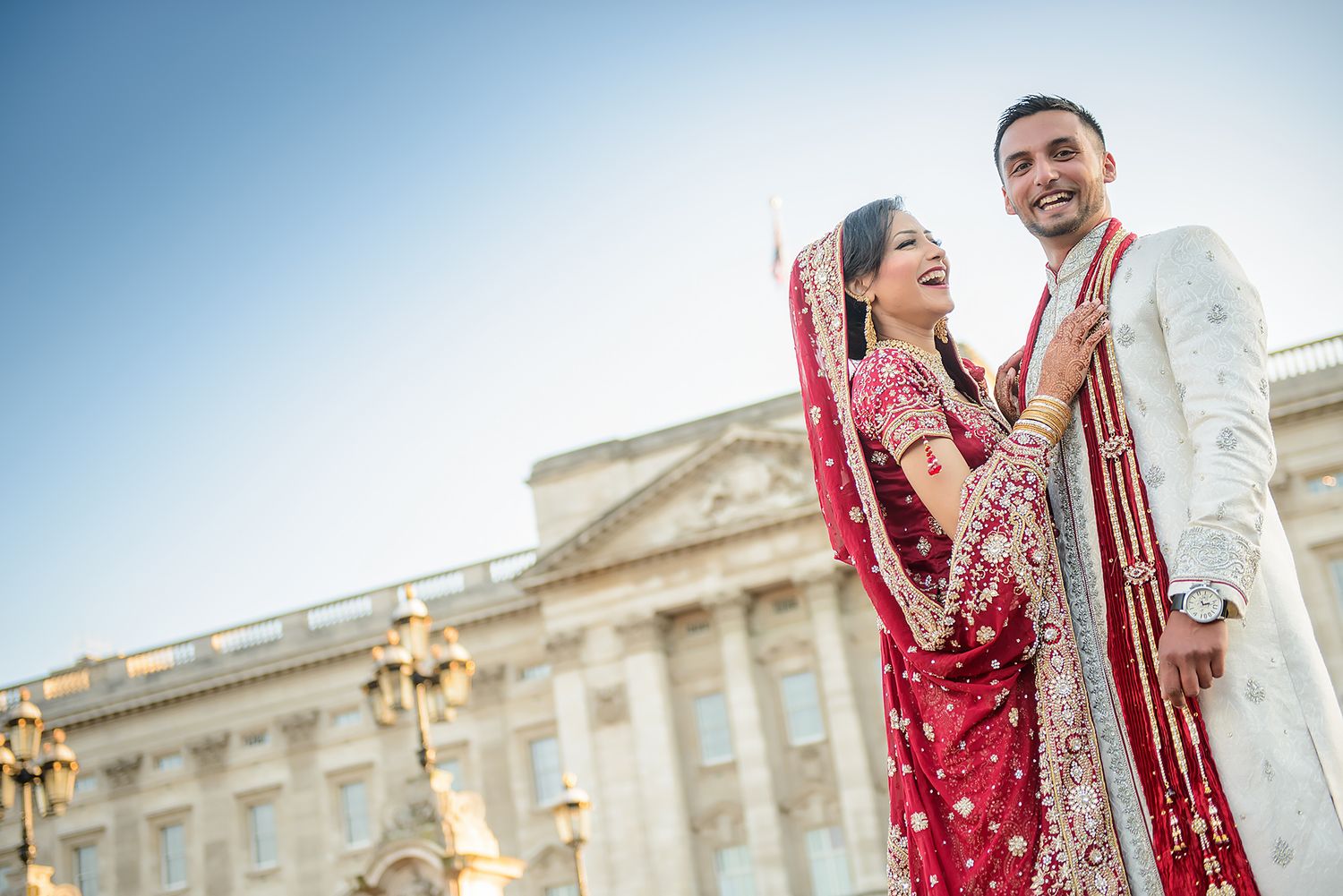 Happy couple in traditional Indian wedding attire poses together outside elegant European building.