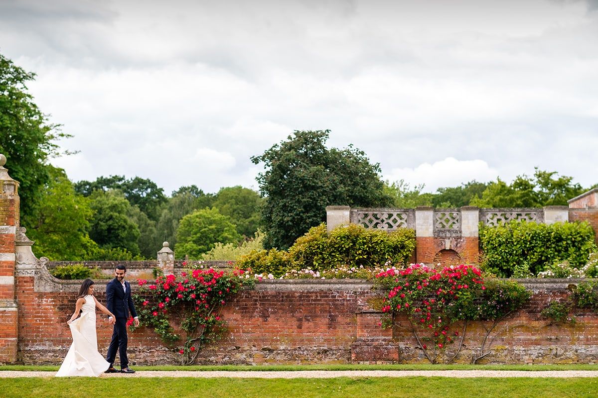 Couple walking along flowering brick wall during their wedding celebration.
