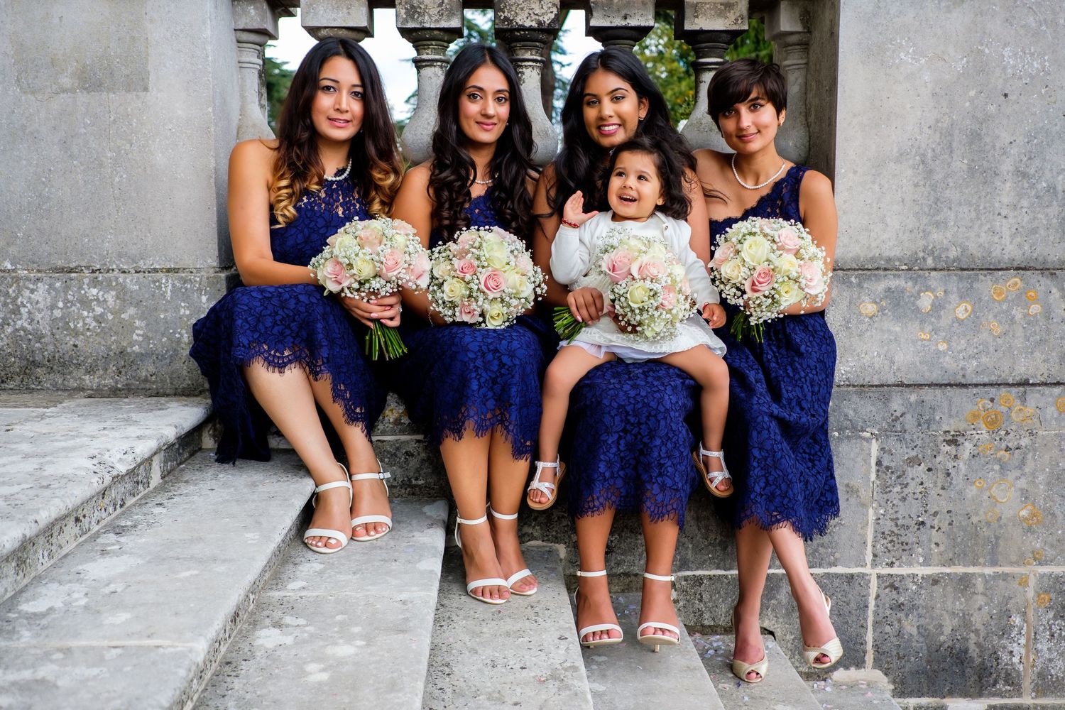 Bridesmaids in blue dresses show off their shoes while seated on stone steps.