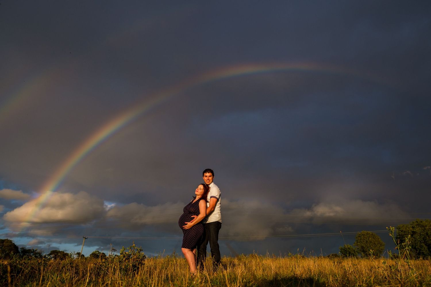 Rainbow arches over golden fields of tall grass against a dramatic cloudy sky during sunset.
