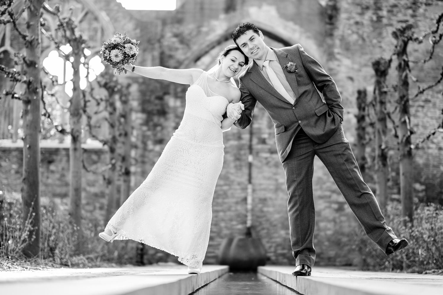 Playful black and white wedding photo of couple striking dramatic pose in garden setting.