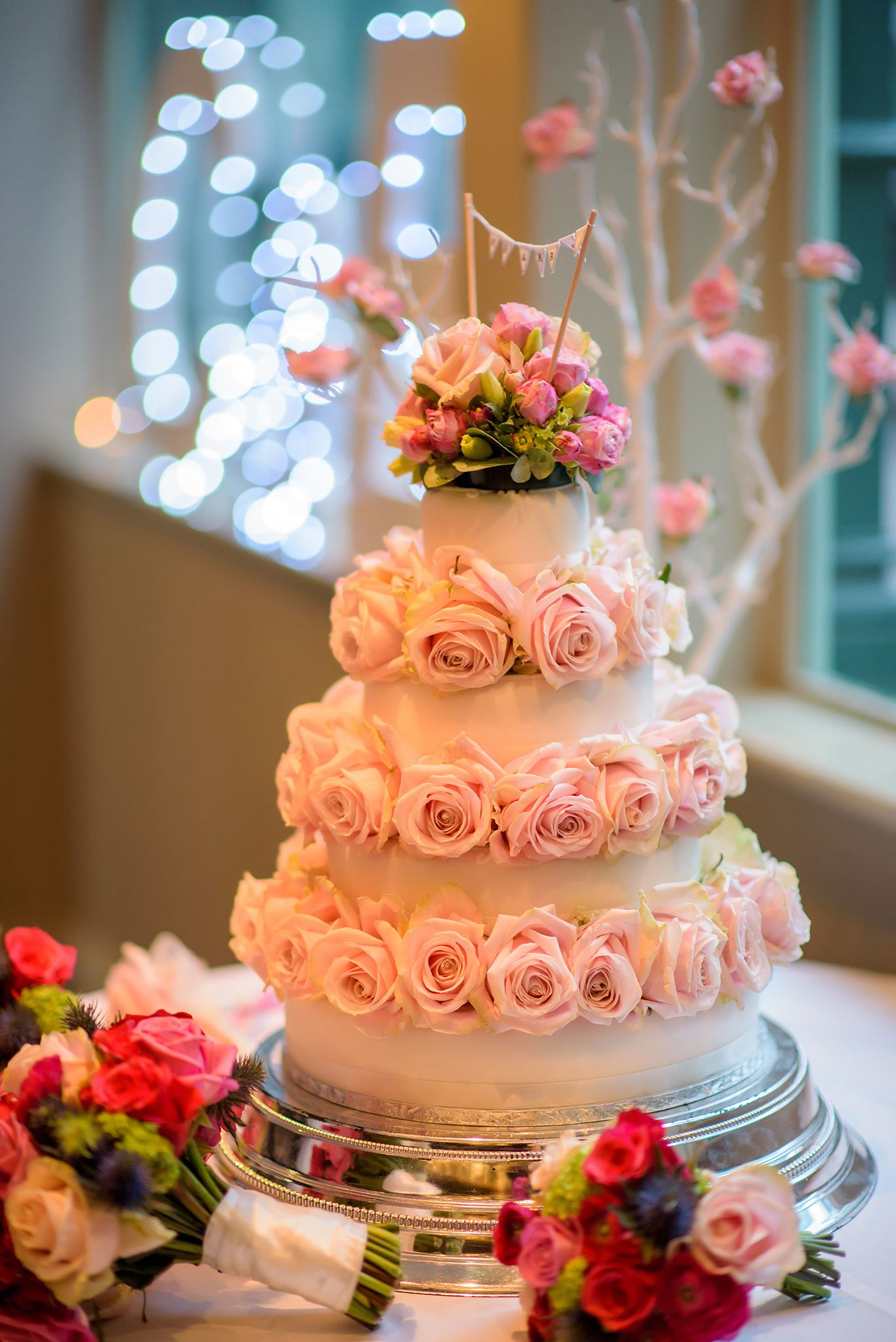 An elegant white wedding cake decorated with pink roses and floral accents against a bokeh background.