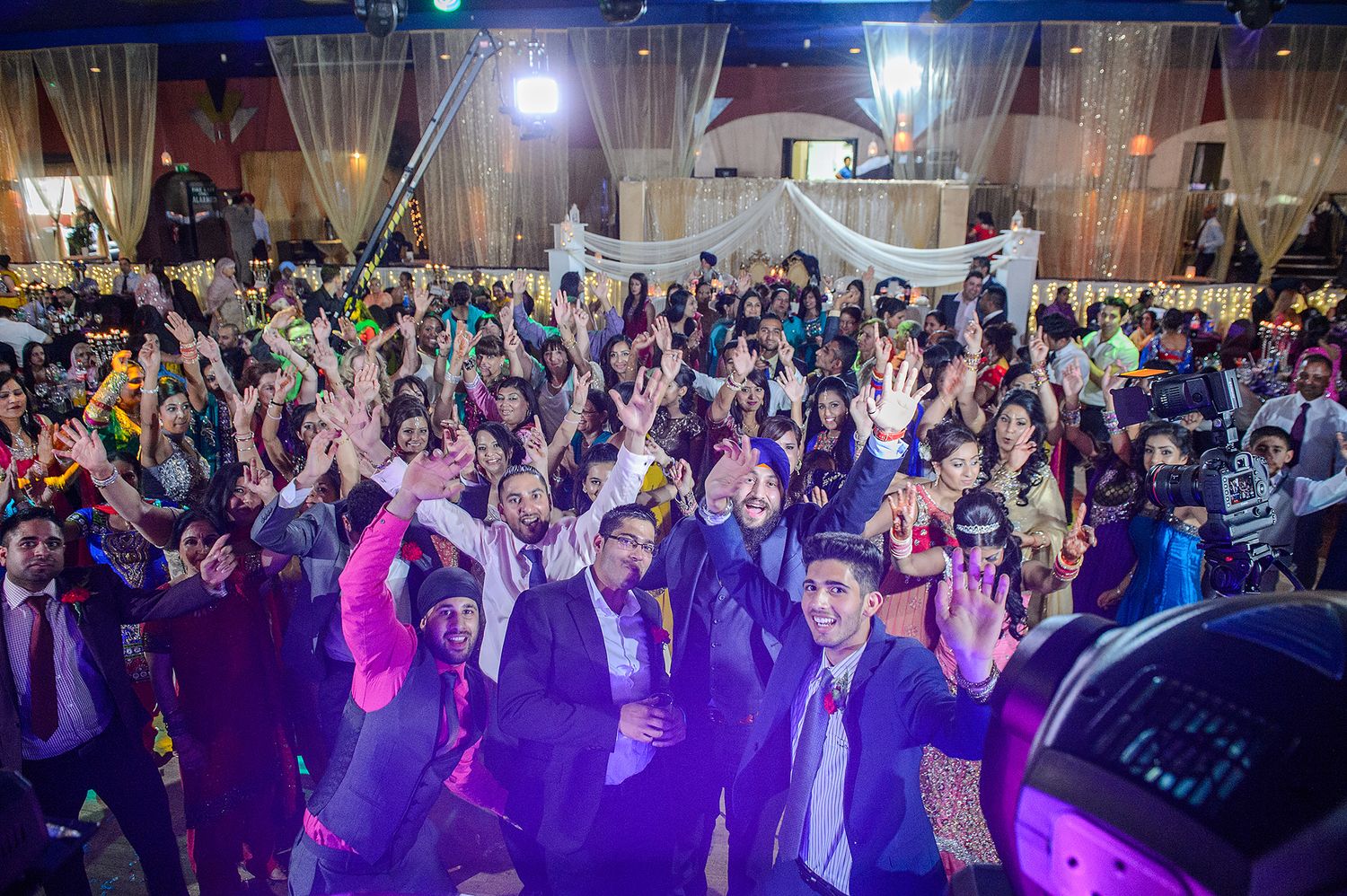 Large crowd dancing and celebrating at a wedding reception with purple mood lighting.